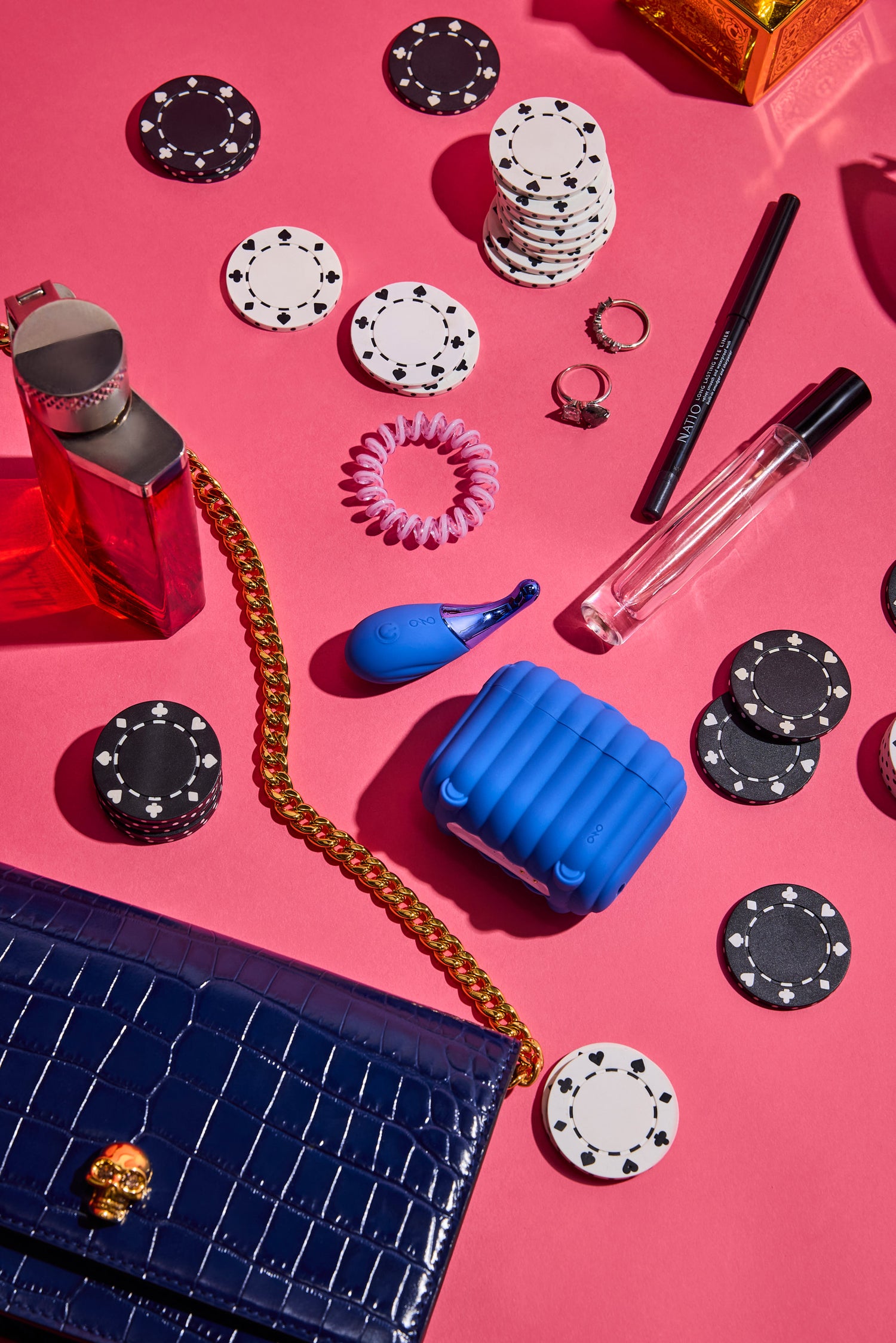 Assorted items including poker chips, a blue inflatable cup, a black handbag, and a pink hair tie on a pink background.